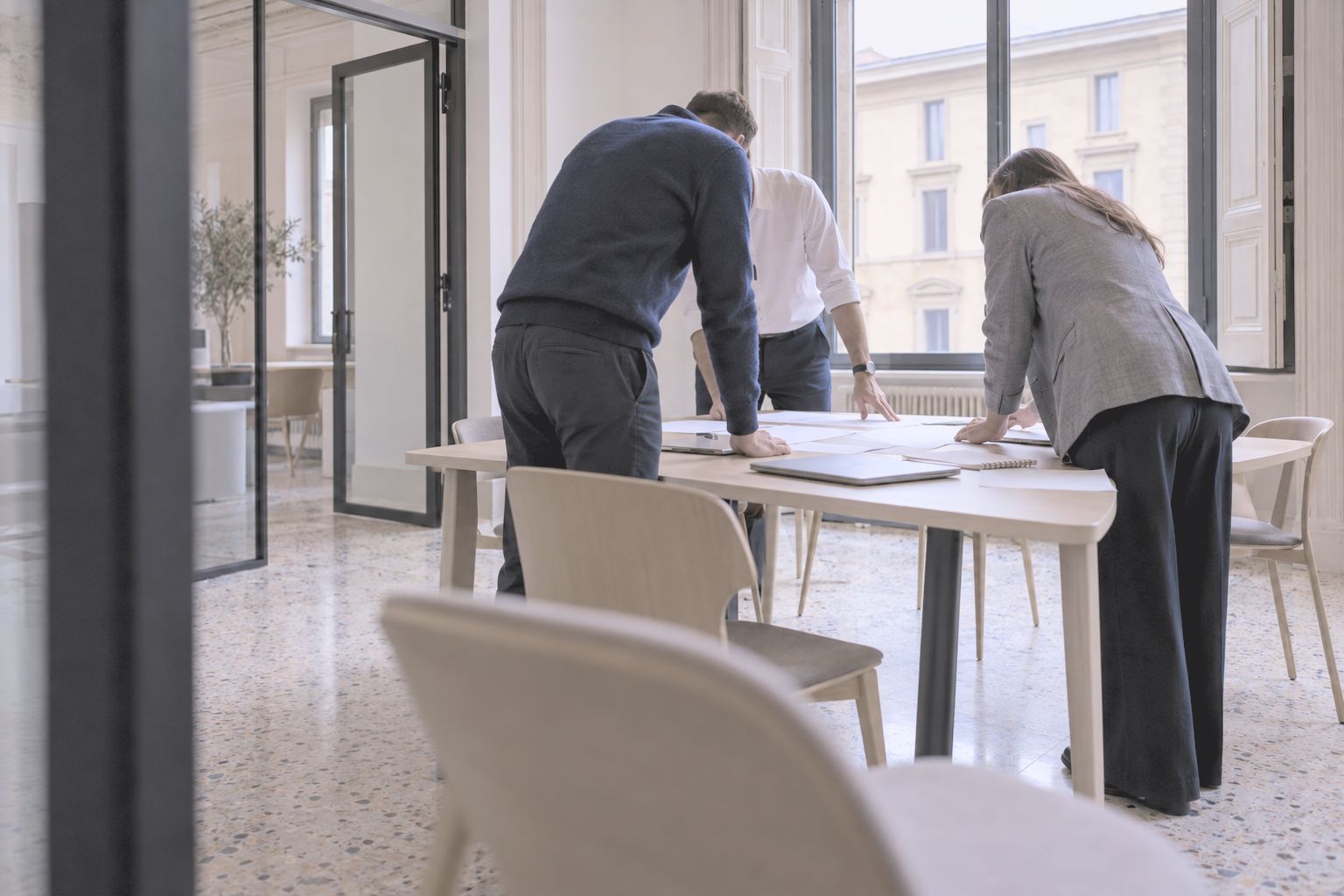 Three colleagues leaning over papers at a table by tall windows